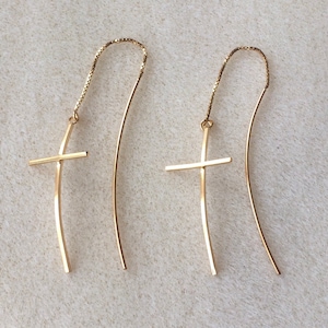 May include: A pair of gold-coloured cross earrings. Each earring has a delicate cross design suspended from a fine chain and curved wire. The earrings are shown against a neutral background.