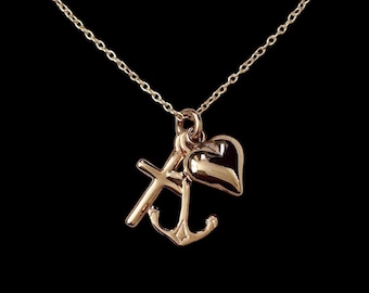 May include: A gold-coloured necklace featuring three charms: a cross, an anchor, and a heart. The chain and charms are gold-toned, set against a black background. The necklace is a pendant style.