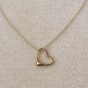 May include: A gold-toned necklace featuring a delicate chain and a single open heart-shaped pendant. The pendant is smooth and minimalist in design, suspended from a thin, polished metal chain that connects to the top of the heart shape.