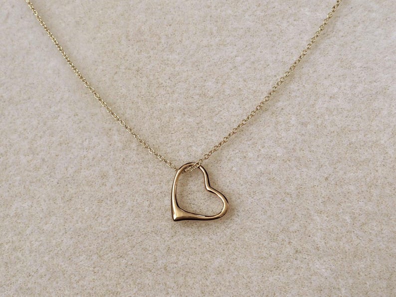 May include: A gold-toned necklace featuring a delicate chain and a single open heart-shaped pendant. The pendant is smooth and minimalist in design, suspended from a thin, polished metal chain that connects to the top of the heart shape.