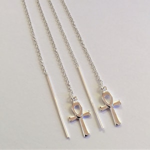 May include: A pair of silver threader earrings with an Ankh symbol pendant. The earrings have a fine chain and a straight bar. The Ankh pendant is a cross with a loop at the top. The earrings are shown against a white backdrop.