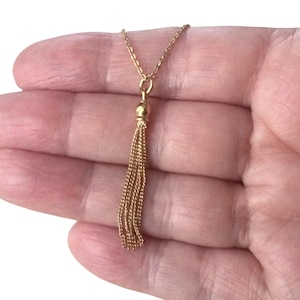 May include: A gold-coloured necklace with a tassel pendant. The necklace chain is delicate, and the tassel is made of multiple strands of gold-coloured chain. The necklace is displayed on a hand.
