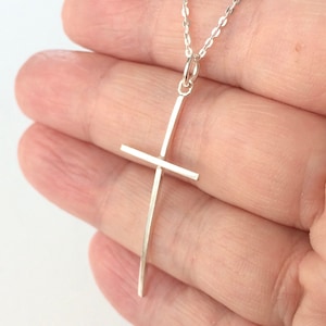 May include: A silver cross pendant necklace. The cross is a simple, minimalist design with a vertical bar and a horizontal bar. The necklace chain is delicate and silver. The pendant is held in a hand.