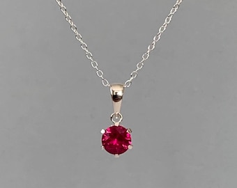 May include: A silver necklace with a small, round, bright red gemstone pendant. The pendant is held in place by four prongs. The chain is delicate and hangs against a gray background.