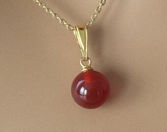 May include: A gold chain necklace with a red carnelian gemstone pendant. The pendant is a round, smooth sphere.