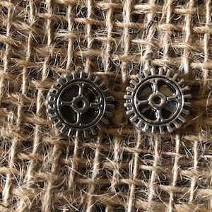 May include: Two silver gear-shaped earrings with a black center. The earrings are on a brown burlap background.