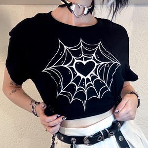 Pode incluir: Camiseta preta com um desenho de teia de aranha branca e um coração no centro. A modelo usa um colar gargantilha preto com um pingente de coração, uma saia plissada branca e um cinto preto. Acessórios: pulseiras e orelhas de gato.