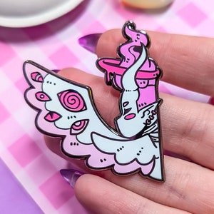 May include: Pink and white enamel pin featuring a winged creature with a candle on its head. The creature has large eyes and a swirling pattern on its wings.