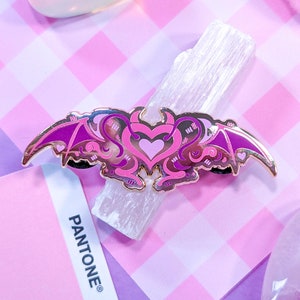 May include: A gold-tone enamel pin with a pink and purple heart design with bat wings. The heart has a small devil horn on top.