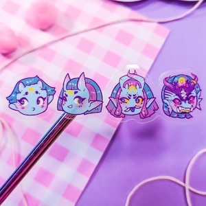 Può includere: Quattro adesivi trasparenti con illustrazioni colorate di personaggi dei cartoni animati con capelli blu e viola e corna. I personaggi hanno diverse espressioni facciali, tra cui un'espressione sorpresa, un'espressione sorridente, un'espressione ammiccante e un'espressione accigliata.