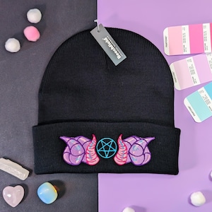 May include: Black beanie with an embroidered design featuring purple and pink horns, a pentagram, and a tag that reads "Beechfield". The hat is displayed on a purple and gray surface with color swatches and decorative items.