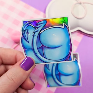 Könnte beinhalten: Zwei Aufkleber mit einer Cartoon-Illustration einer blauen Figur mit abgerundeter Form. Die Aufkleber haben einen holografischen Rand und werden von einer Hand mit lila Nagellack gehalten. Der Hintergrund ist eine Mischung aus Rosa und Lila.