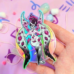 May include: A holographic sticker featuring a pink and white angel with multiple eyes, wings, and a halo. The angel is standing on a pentagram inside a circle with a rainbow gradient.