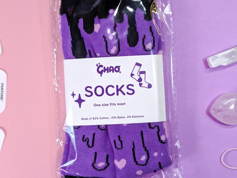 May include: Purple and black socks with a dripping design and hearts. The socks are packaged in a clear plastic bag with a white label that reads "CHAO SOCKS One size fits most Made of 82% Cotton, 15% Nylon, 3% Elastane".