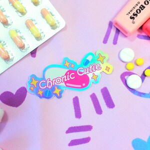 May include: A holographic sticker with a pink pill shape and the text "Chronic Cutie" in white letters. The sticker is surrounded by yellow stars and is on a pink background with purple and blue hearts and lines.