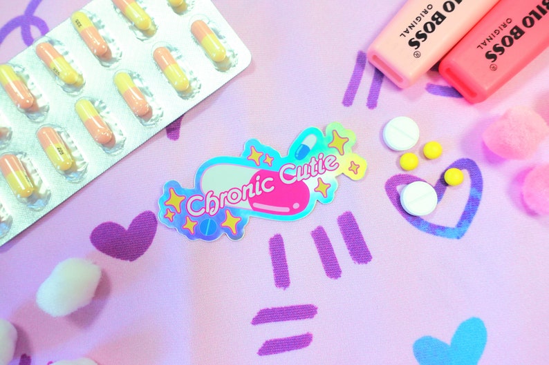 May include: A holographic sticker with a pink pill shape and the text "Chronic Cutie" in white letters. The sticker is surrounded by yellow stars and is on a pink background with purple and blue hearts and lines.