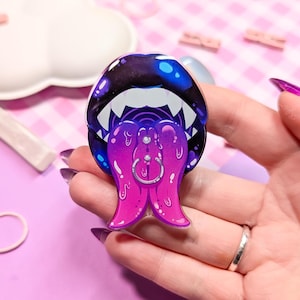 May include: A black and purple enamel pin depicting a mouth with a large, dripping tongue. The tongue is pink and purple and has a silver ring piercing through it.
