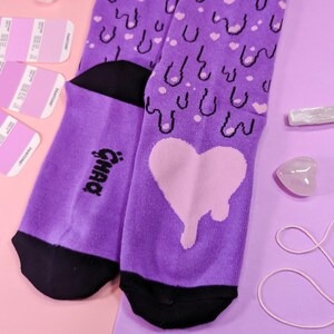 May include: Purple socks with a dripping heart and a repeating pattern of hearts and drips. The socks have a black heel and toe. The text "Gnar" is printed on the side of the sock.