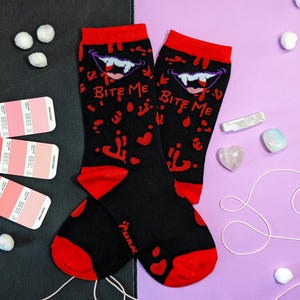 May include: A pair of black socks with red accents. The socks feature a white vampire mouth with the text "Bite Me" on each sock. The socks are on a purple background with white and blue crystals.