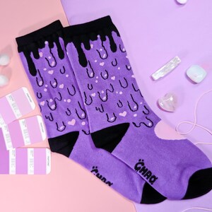 May include: Purple socks with a black dripping design and pink hearts. The socks have the word "GHAQ" written on them.