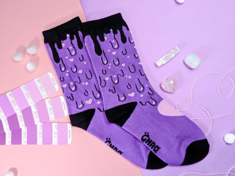 May include: Purple socks with a black dripping design and pink hearts. The socks have the word "GHAQ" written on them.