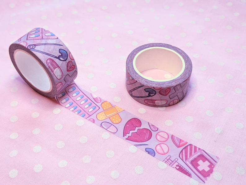 May include: Two rolls of washi tape with a pink and white polka dot background. The tape features a pattern of medical supplies, including bandages, pills, and a heart.