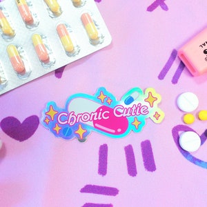 May include: A holographic sticker with a pink pill shape and the text "Chronic Cutie" in white letters. The sticker is surrounded by yellow stars and is on a pink background with purple lines and hearts.