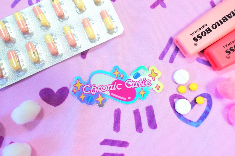 May include: A holographic sticker with a pink pill shape and the text "Chronic Cutie" in white letters. The sticker is surrounded by yellow stars and is on a pink background with purple lines and hearts.