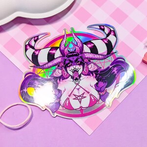 May include: A holographic sticker featuring a purple demon with black and white horns, a pentagram, and a rainbow. The demon is wearing a pink bikini top with a pentagram.