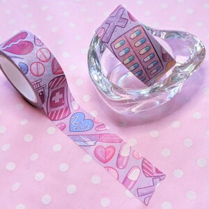 May include: Two rolls of washi tape with a pink and white polka dot background. One roll features a pink and white design with hearts, pills, and syringes. The other roll features a pink and white design with pills and crosses.