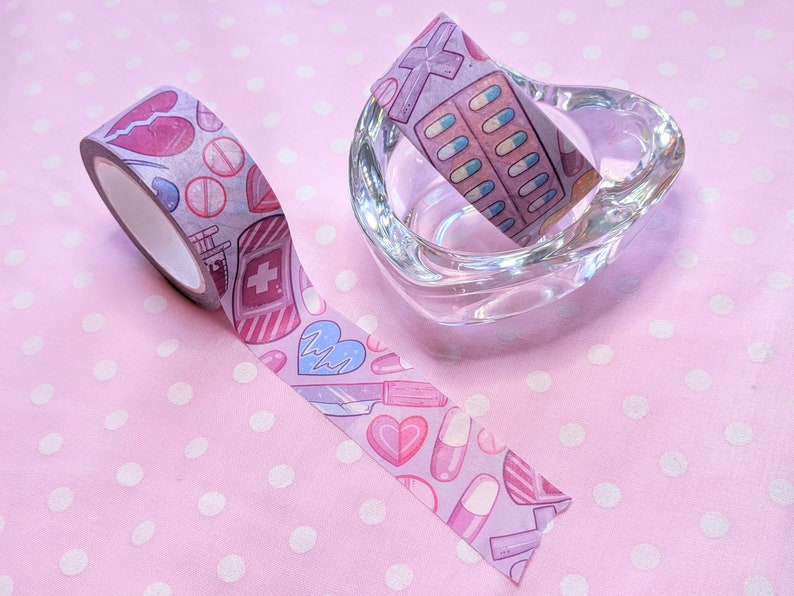 May include: Two rolls of washi tape with a pink and white polka dot background. One roll features a pink and white design with hearts, pills, and syringes. The other roll features a pink and white design with pills and crosses.