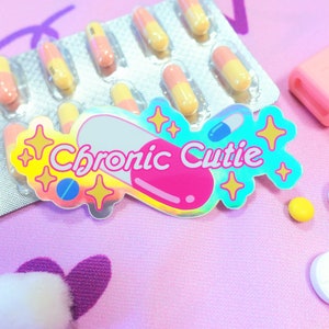 May include: A holographic sticker with the text "Chronic Cutie" in a pink and blue pill shape with gold stars. The sticker is on a pink surface with a pill container, white pills, and a pink highlighter.