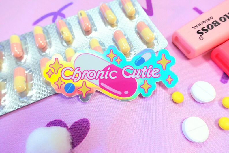 May include: A holographic sticker with the text "Chronic Cutie" in a pink and blue pill shape with gold stars. The sticker is on a pink surface with a pill container, white pills, and a pink highlighter.