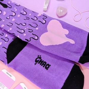 May include: Purple and black socks with a pink heart and dripping black paint design. The socks have the word "GHAD" written on them.