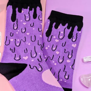 May include: A pair of purple socks with a black dripping design and pink hearts. The socks are on a pink and purple background.