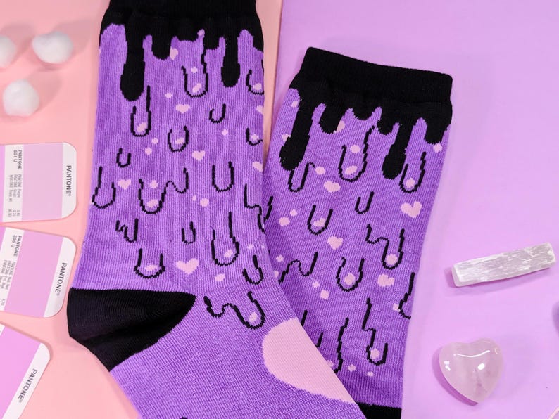 May include: A pair of purple socks with a black dripping design and pink hearts. The socks are on a pink and purple background.