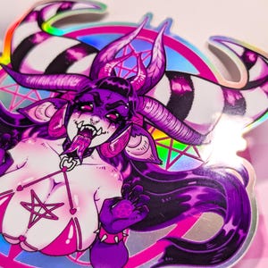 May include: A holographic sticker featuring a purple and pink demon with horns, a pentagram, and a pink bikini. The demon has a long tongue sticking out and is surrounded by a rainbow halo.
