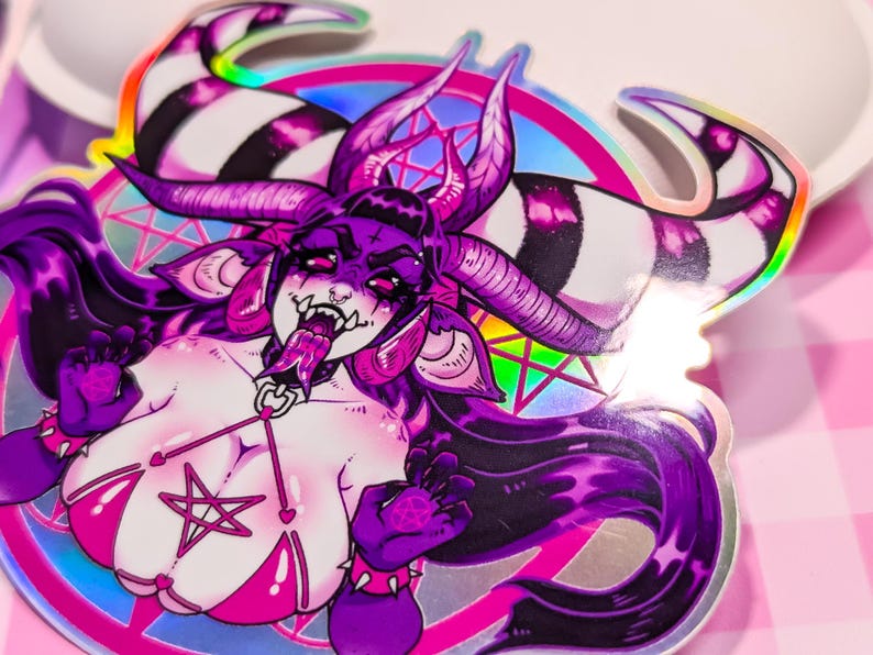 May include: A holographic sticker featuring a purple and pink demon with horns, a pentagram, and a pink bikini. The demon has a long tongue sticking out and is surrounded by a rainbow halo.