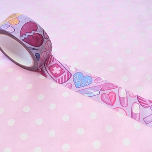 May include: Pink washi tape with a pattern of hearts, pills, bandages, and a scalpel. The tape is rolled up and partially unrolled on a pink background with white polka dots.