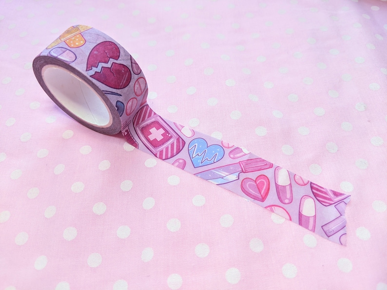 May include: Pink washi tape with a pattern of hearts, pills, bandages, and a scalpel. The tape is rolled up and partially unrolled on a pink background with white polka dots.