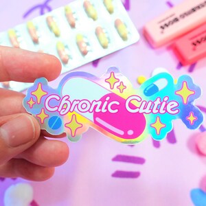 May include: A holographic sticker with a pink pill design and the text "Chronic Cutie". The sticker has a rainbow iridescent finish and features glitter stars.