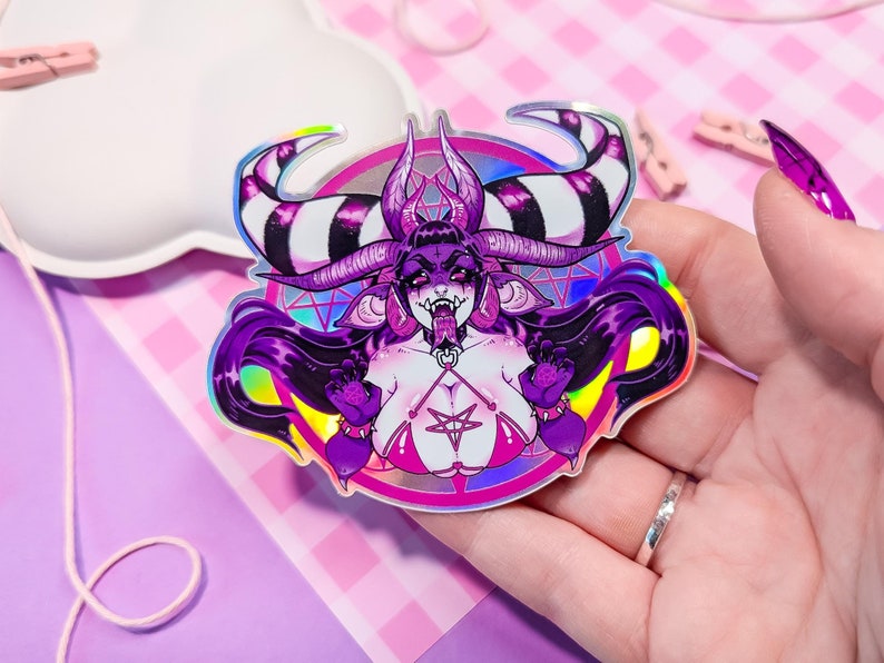 May include: A holographic sticker featuring a purple demon with horns, long black hair, and a pentagram symbol. The demon is wearing a pink and black bikini and a choker with a pentagram pendant. The sticker has a rainbow iridescent finish.