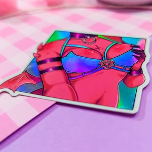 Może przedstawiać: Holograficzna naklejka przedstawiająca czerwonosk&oacute;rą postać z rogami i skrzydłami, ubraną w niebieski i fioletowy, tęczowy top bikini z pentagramem. Postać ma czerwoną i czarną obrożę z srebrnym pierścieniem.