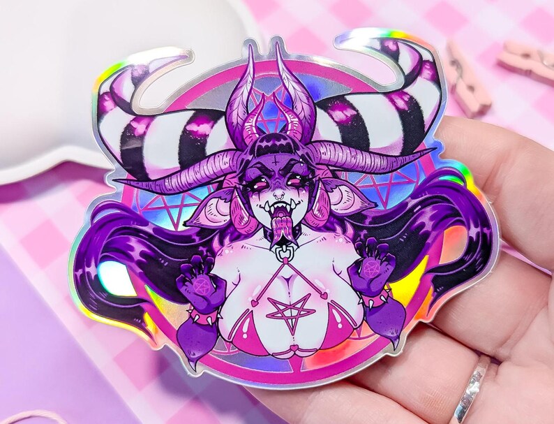 May include: A holographic sticker featuring a purple and pink demon with black and white striped horns, wearing a pink bikini top with a pentagram. The demon has long purple hair and is surrounded by a pink and purple circle with a pentagram.