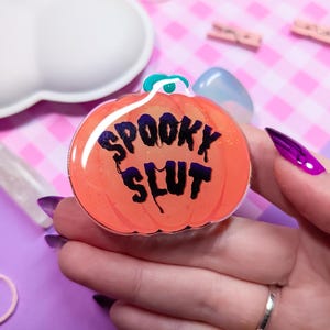 May include: Orange pumpkin-shaped resin pin with the text "SPOOKY SLUT" in black lettering.
