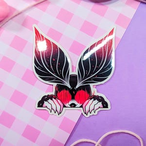 May include: A sticker featuring a black and red creature with large, leafy ears peeking out from behind a wall. The creature has red eyes and sharp teeth.