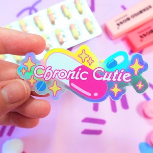 May include: A holographic sticker with a pink and white pill design. The sticker says "Chronic Cutie" with a blue and white pill inside the pill design. The sticker is surrounded by yellow stars.
