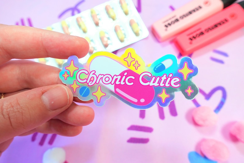 May include: A holographic sticker with a pink and white pill design. The sticker says "Chronic Cutie" with a blue and white pill inside the pill design. The sticker is surrounded by yellow stars.