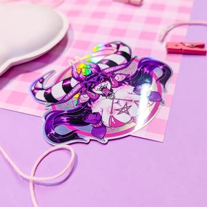 May include: A holographic sticker featuring a purple and white demon character with horns, a pentagram, and a pink background.