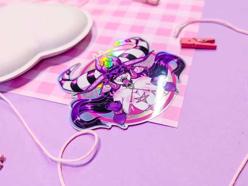 May include: A holographic sticker featuring a purple and white demon character with horns, a pentagram, and a pink background.